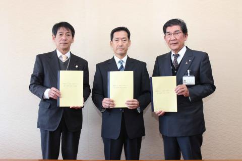 スーツを着た男性が3人横一列に並び合意書を持ってる締結式写真